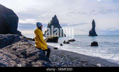 Un voyageur est assis sur le sable noir de Reynisfjara Beach Islande, contemplant les majestueuses piles de mer s'élevant de l'océan sous un ciel nuageux. Les vagues calmes tournent doucement sur le rivage Banque D'Images