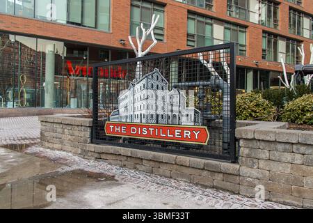 Panneau Distillery District devant un bâtiment en briques avec aménagement paysager hivernal. Un quartier historique connu pour son architecture victorienne et son charme saisonnier. Banque D'Images