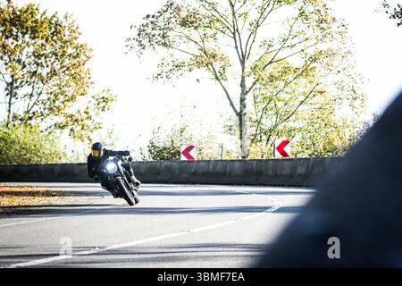 Motocycliste prenant un virage serré sur une route pittoresque entourée d'arbres d'automne, capturé en mouvement avec des couleurs saisonnières vibrantes. Banque D'Images