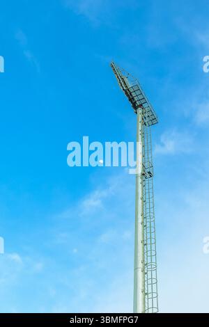 Poteau de projecteur de stade à faible angle contre le ciel bleu avec des nuages et un petit croissant de lune. Infrastructure sportive moderne, vue verticale. Banque D'Images