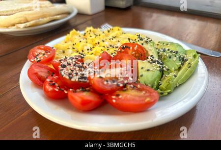 Gros plan d'un petit déjeuner sain servi dans un café ou un restaurant : omelette, tomates tranchées et avocat nappé de graines sur une assiette, sur une table en bois. Banque D'Images