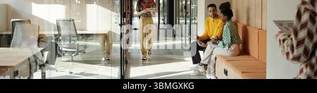 Homme noir et femme noire assis sur le banc orange discutant des documents dans l'espace de coworking moderne tandis que la jeune femme avec l'ordinateur portable marche Banque D'Images