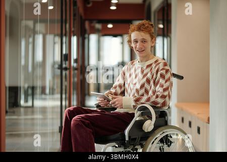 Jeune homme caucasien adulte avec les cheveux roux assis en fauteuil roulant tenant une tablette numérique et souriant, portant un pull à motifs et des écouteurs Banque D'Images