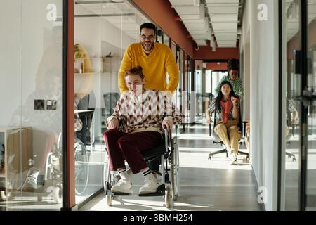 Jeune homme noir poussant l'homme caucasien avec les cheveux roux en fauteuil roulant à travers le bureau moderne, de jeunes collègues féminines s'amusant en arrière-plan Banque D'Images