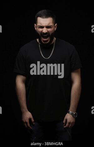 Portrait dramatique d'un jeune homme sérieux, musicien, chanteur, rappeur avec une barbe en vêtements noirs sur un fond noir isolé. il crie de colère Banque D'Images