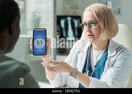 Médecin féminin d'âge moyen caucasien montrant smartphone avec application de niveau de glucose au patient masculin d'âge moyen noir pendant la consultation de diabète en médecine Banque D'Images