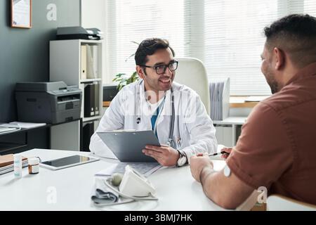 Médecin masculin caucasien d'âge moyen souriant tout en consultant un homme hispanique d'âge moyen atteint de diabète dans un cabinet médical, patient assis au bureau avec gluc Banque D'Images