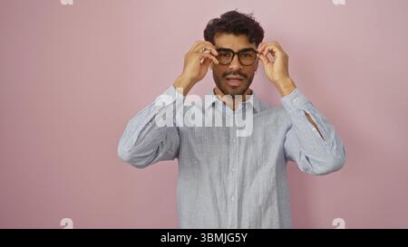 Jeune homme avec la barbe ajuste les lunettes tout en se tenant debout contre un mur rose, exsudant la confiance et le style dans une chemise décontractée, incarnant moderne attrayant m Banque D'Images