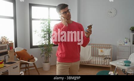 Jeune homme en pull rouge parle au téléphone dans une chambre bien éclairée tenant bouteille de médecine près du lit de bébé avec des jouets, des plantes et une horloge visible dans l'indoo confortable Banque D'Images