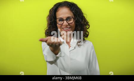 Femme hispanique aux cheveux bouclés étend sa main vers la caméra sur un fond jaune vif, exsudant la confiance et l'accessibilité. Banque D'Images