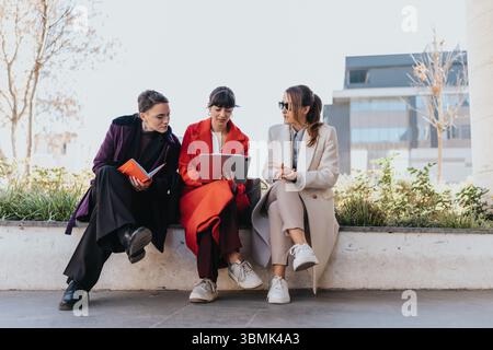 Trois femmes d'affaires collaborant à l'extérieur examinant un ordinateur portable tout en étant assis sur un banc dans un cadre urbain moderne Banque D'Images