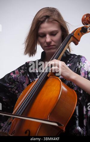 Jeune femme caucasienne adulte jouant du violoncelle en studio, portant une robe de satin floral. Gros plan. Banque D'Images