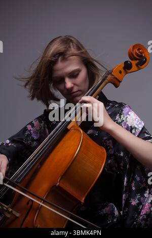 Gros plan en studio d'une jeune femme caucasienne adulte jouant du violoncelle. Elle porte une robe noire avec un motif floral violet et rose et a un f Banque D'Images