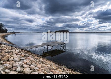 Paisible vue sur la mer mettant en valeur une jetée en bois s'étendant dans des eaux calmes, encadrée par des rochers et un ciel bleu vif avec des nuages, évoquant la sérénité et le natu Banque D'Images