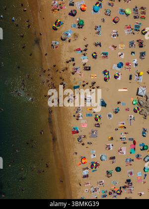 Drone aérien vue de dessus en bas d'une plage bondée avec des parasols colorés et des bains de soleil pendant un après-midi d'été près de la côte - capturé à Cascais Banque D'Images