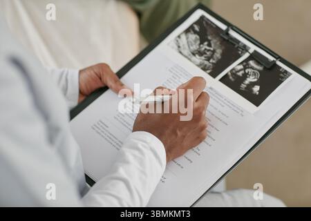 Gros plan de femme noire tenant un stylo et écrivant sur un formulaire médical avec des images échographiques attachées au presse-papiers, documentant les soins prénataux pour une femme enceinte pendant la visite de contrôle Banque D'Images