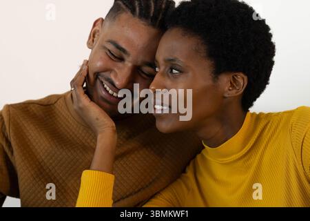 Couple romantique souriant penchant les têtes ensemble contre le mur léger, avec détail anneau or Banque D'Images