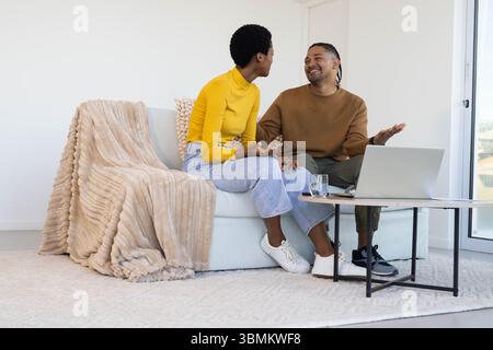 Parler couple diversifié assis sur le canapé dans le salon moderne, avec ordinateur portable et verre d'eau Banque D'Images