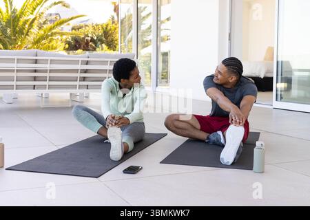 Étirer un couple diversifié en vêtements de sport faisant des étirements de jambe sur un patio carrelé avec des tapis de yoga et un téléphone Banque D'Images