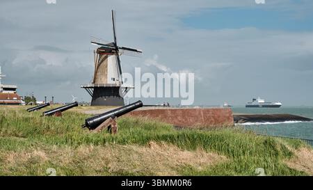 28 mai 2025 - Vlissingen-pays-Bas : moulin à vent hollandais traditionnel sur la digue de la Westerschelde, vieux canons devant, éoliennes et shippin Banque D'Images