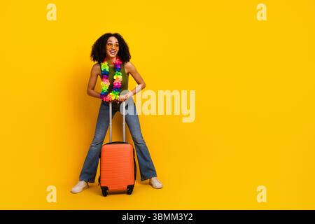 Jeune femme colorée avec valise orange et lei hawaïen sur fond jaune vif embrassant des vibrations de voyage Banque D'Images