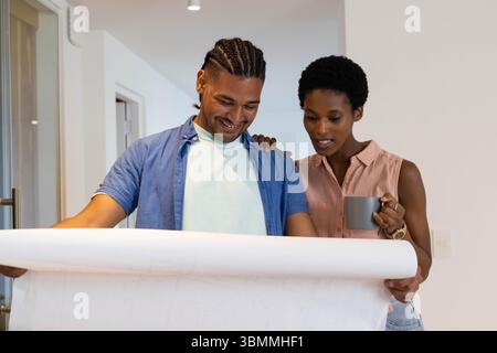 Examen du couple diversifié debout dans le couloir moderne regardant un grand plan, avec tasse en céramique Banque D'Images