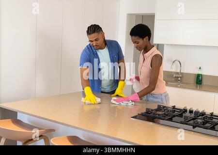 Essuyer le comptoir d'îlot en pierre dans la cuisine, couple diversifié portant des gants en caoutchouc à l'aide d'un vaporisateur Banque D'Images