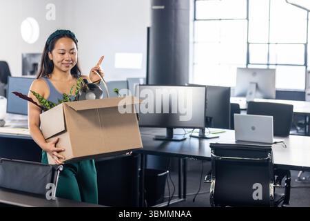 Femme chinoise marchant à travers le bureau avec boîte mobile contenant la plante en pot, lampe de bureau, espace de copie Banque D'Images