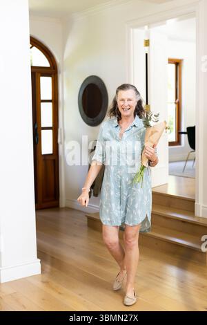 Femme senior se promenant dans le foyer tenant la verdure et bouquet de fleurs blanches près de la porte en bois voûtée Banque D'Images