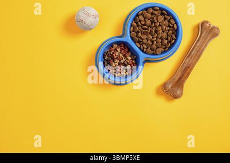 Séchez la nourriture pour animaux dans un bol avec une boule et un os pour chiens sur fond jaune vue du dessus. Encore la vie. Copier l'espace. Pose à plat Banque D'Images