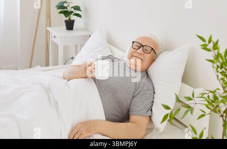 Homme âgé senior allongé sur le lit dans la chambre tenant une tasse de café ou de thé appréciant bonjour Banque D'Images