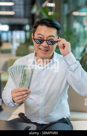 Heureux riche homme d'affaires chinois mûr portant des lunettes de soleil Thug Life montrant des billets de banque d'argent dollar assis dans le hall moderne du bureau. Carrière d'affaires de succès, gagnant de loterie, grand revenu, richesse. Banque D'Images