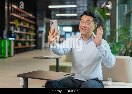Portrait de jeune homme d'affaires souriant tenant smartphone agitant le signe de main bonjour saluant interlocuteur dans la conférence en ligne appel vidéo à distance du hall du bureau. Un gars en vêtements formels assis dans le hall. Banque D'Images