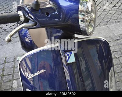 Vespa bleu brillant garé sur une rue pavée, reflétant les bâtiments environnants Banque D'Images