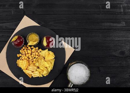 Vue en hauteur sur un verre à bière et des en-cas avec sauce sur une table en bois noir. Vue de dessus. Copier l'espace. Encore la vie. Pose à plat Banque D'Images