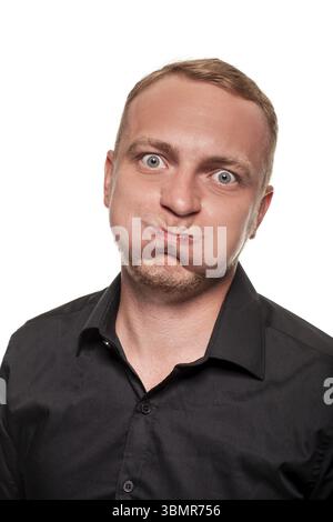 Beau jeune homme blond dans une chemise noire fait des visages et gonfle ses joues, isolé sur un fond blanc Banque D'Images
