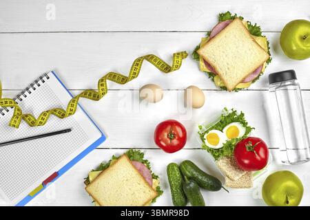 Vue de dessus de l'ordinateur portable de planification avec copie espace et petit déjeuner sain. Sandwich au jambon et fromage, œufs, le concombre, la tomate et une bouteille d'eau sur blanc wo Banque D'Images