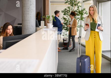 Femme d'affaires confiante avec valise et passeport à l'aide d'un smartphone tout en se tenant debout à la réception de l'hôtel Banque D'Images