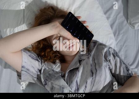 Une jeune femme est allongée sur son lit avec une expression peinte, couvrant son front d'une main. Elle regarde son téléphone alors que la lumière du soleil filtre à travers les rideaux, créant une atmosphère calme mais tendue. Banque D'Images