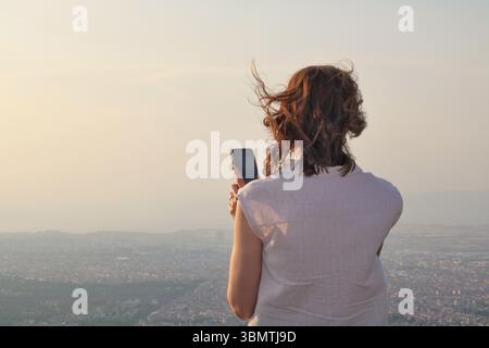 Femme prenant une photo du paysage urbain au coucher du soleil avec son téléphone depuis un panorama, vent dans ses cheveux. Banque D'Images