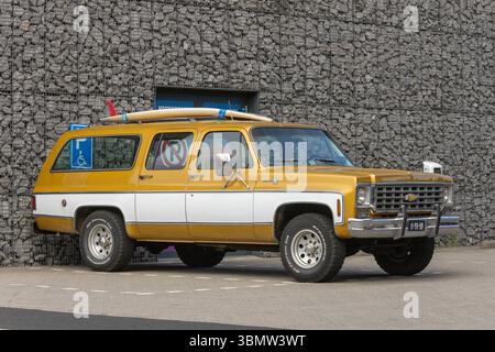 VUS Chevrolet Suburban vintage garé devant un mur de pierre moderne, avec planches de surf sur le toit et peinture rétro audacieuse or et blanc. Leiden, N. Banque D'Images