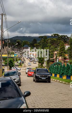 Rue pavée animée dans la station de montagne Monte Verde avec des voitures venant en sens inverse, des magasins et des arbres araucaria sous un ciel nuageux spectaculaire. Banque D'Images