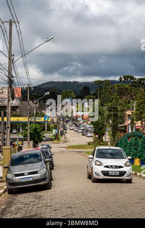 Rue pavée animée dans la station de montagne Monte Verde avec des voitures, des magasins et des arbres araucaria sous un ciel orageux spectaculaire à Monte Verde. Banque D'Images
