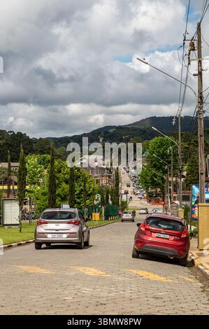Rue principale pavée (LMG-886) de la station de montagne Monte Verde avec des voitures, des cyprès et des collines lointaines sous le ciel nuageux. Banque D'Images