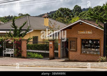 Façade en brique de la boutique Essências de Minas avec banc de jardin et portail sur l'avenue Monte Verde, sous ciel nuageux avec des pins à l'arrière. Banque D'Images