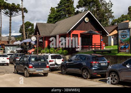 Garagem Vemaguet bar bâtiment rouge de style alpin avec des voitures garées sur l'avenue Monte Verde, encadré par des arbres d'araucaria sous ciel nuageux. Banque D'Images