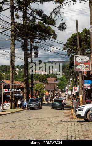 Rue principale pavée animée de la station de montagne Monte Verde bordée d'arbres d'araucaria, de magasins, de voitures et de piétons sous le ciel nuageux. Banque D'Images