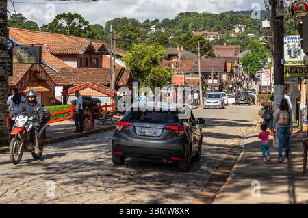 Scène de rue pavée occupée sur l'avenue Monte Verde, avec des voitures, moto, piétons, magasins, et araucaria arbres sous ciel nuageux. Banque D'Images