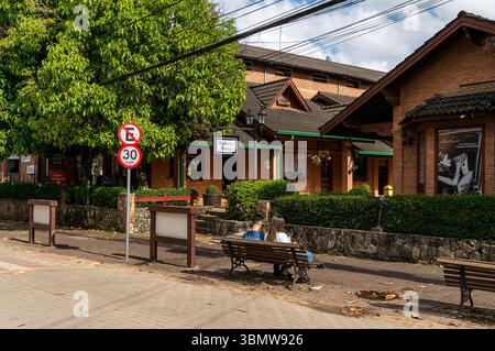 Couple est assis sur un banc en bois face au complexe de construction de briques Galeria Suiça à l'avenue Monte Verde, sous des arbres feuillus et un ciel partiellement nuageux. Banque D'Images