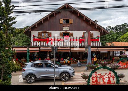 Silver SUV passe devant le grand bâtiment alpin Pousada Avenida inn orné d'arcs de Noël rouges sur l'avenue Monte Verde, sous le ciel nuageux. Banque D'Images